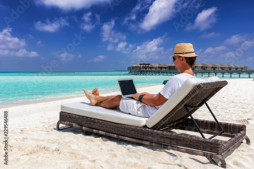 Arbeit im Urlaub: Geschäftsmann mit Laptop auf Liege am tropischen Strand