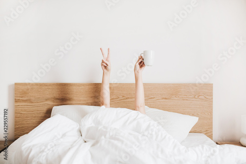 Hand's of young woman with coffee mug in bed with white linens. Minimal happy morning concept.