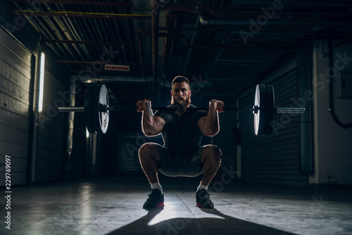 Muscular attractive caucasian bearded man doing front barbell squat in underground hallway. Storage units and pipes in background.