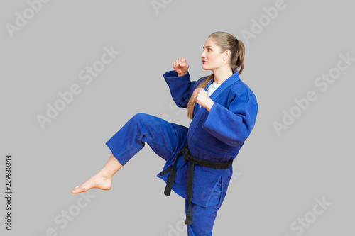 Side view of agressive athletic karate woman in blue kimono with black belt are ready to fight, punching with foot and looking away. Japanese martial arts concept. Indoor, studio shot, gray background