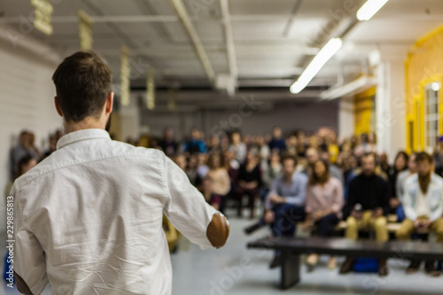 Man with white shirt is giving a conference in front of 200 people in an industrial environment with yellow and white walls - Blurred audience mainly composed of young adults