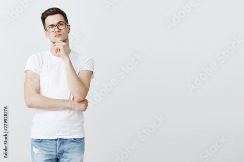 Thoughtful serious-looking male coworker thinkings, trying make up idea, touching chin raising one eyebrow in interest making assumptions or choice, stuck with decision over gray background