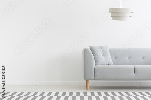 Lamp above grey couch in white minimal apartment interior with copy space on empty wall. Real photo