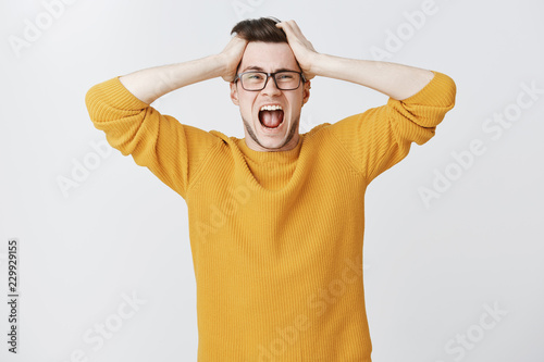 Man yelling cannot control panic and stress holding hands on head open mouth and screaming out loud from distress and anxiety standing troubled and upset in trendy yellow sweater over white background