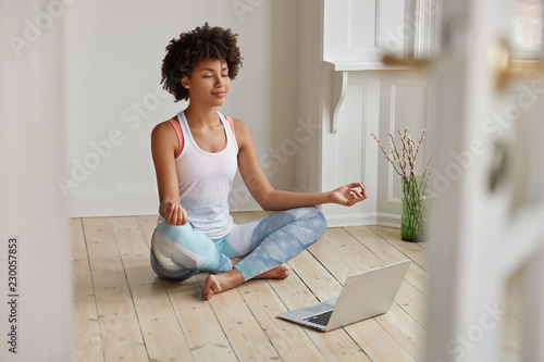 Relaxed dark skinned woman with sporty body, sits in zen pose, keeps legs crossed, watches yoga lessons on laptop computer using internet, meditates on floor in empty room. Wellness concept.