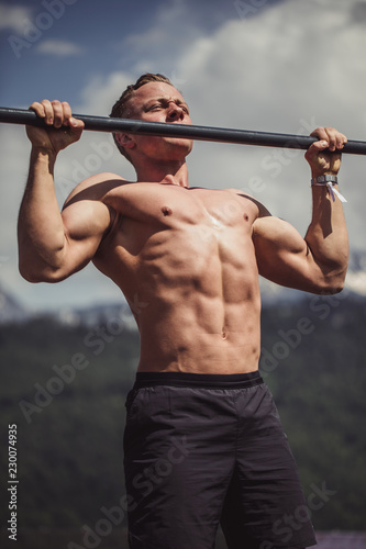 Portrait of a motivated shirtless sportsman doing pull-ups exercise on horizontal bar outdoors, over amazing sky and summer nature background. Sport, health, fitness and urban athletics.