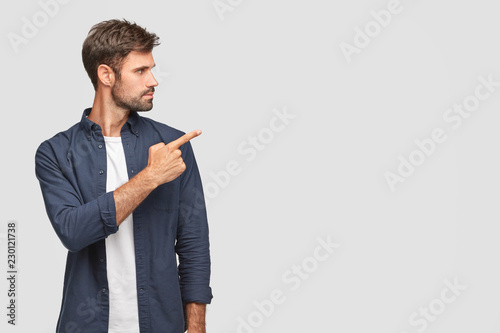 Stylish unshaven young man with dark bristle, attracts your attention to something, points with index finger at blank space, shows free space for your advertising content, stands sideways indoor
