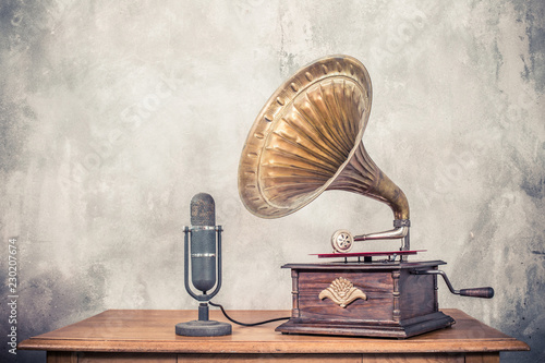 Vintage antique gramophone phonograph turntable with brass horn and big aged studio microphone on wooden table front concrete wall background. Retro old style filtered photo