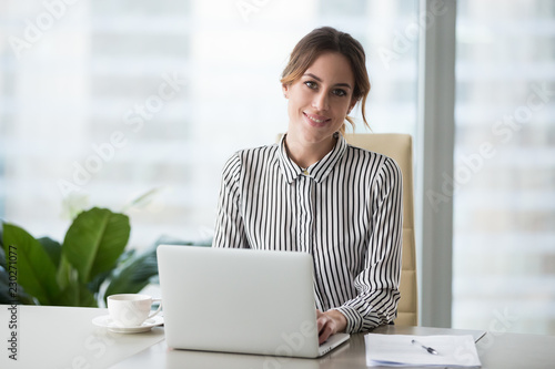 Head shot portrait of confident businesswoman at workplace, smiling woman employee sitting behind laptop and looking at camera. Staff at work.