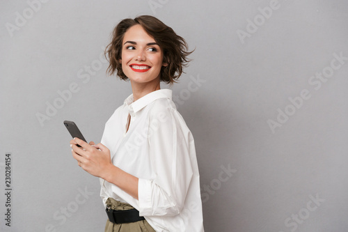 Photo of attractive woman 20s smiling and holding mobile phone, isolated over gray background