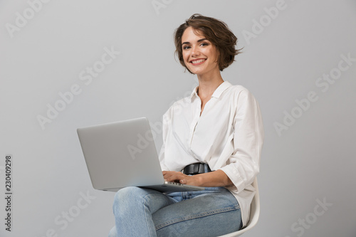 Young business woman posing isolated over grey wall background sitting on stool using laptop computer.