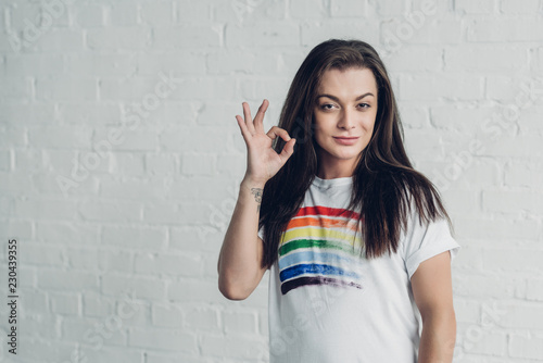 young transgender woman in white t-shirt with pride flag showing okay sign in front of white brick wall