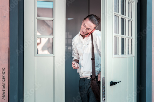 Leaving home. Handsome dark-haired businessman speaking by the phone while leaving home heading to work