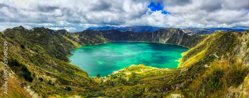 A bird's eye panoramic view of the bright green volcanic Quilotoa Lake in Ecuador with lots of white and grey clouds in a blue sky and green brush on the side of the caldera