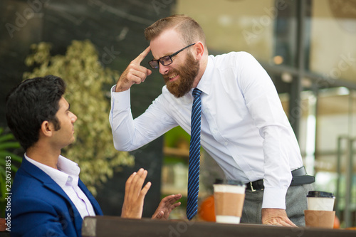 Unstable bearded boss gesturing at head while screaming at Indian employee. Annoyed businessman blaming him for problem, young man taking back and gesturing hands. Aggression concept