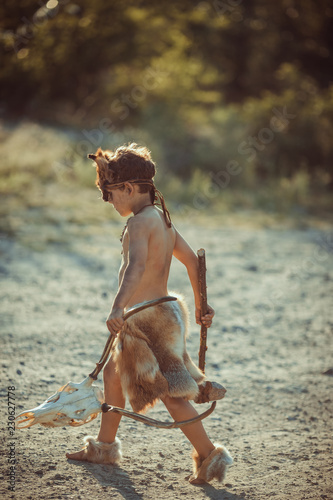 Angry caveman, manly boy with stone axe and animal skull. Prehistoric tribal boy outdoors on nature. Young shaggy and dirty savage, warrior and hunter with weapon. Primitive ice age man in animal skin