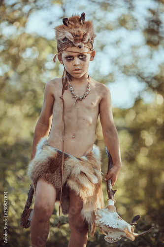 Angry caveman, manly boy with stone axe and animal skull. Prehistoric tribal boy outdoors on nature. Young shaggy and dirty savage, warrior and hunter with weapon. Primitive ice age man in animal skin