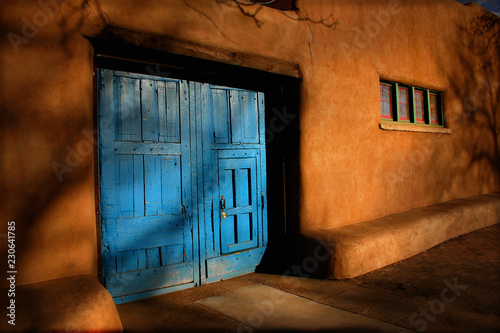 Bright blue wooden doors and Adobe walls of this Santa Fe, New Mexico street view with shadows of trees and natural light of this high altitude plateau.