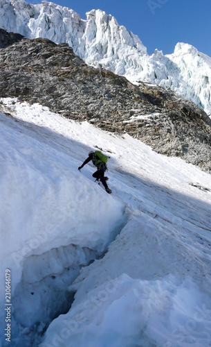 mountain climber crosses over a large and deep crevasse or bergschrund as he begins his climb of a steep north face of ice and snow in the Alps