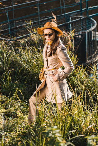 stylish female model in sunglasses, trench coat and hat posing near grass