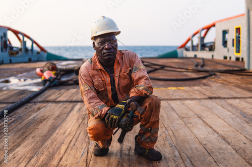 Seaman AB or Bosun on deck of offshore vessel or ship , wearing PPE personal protective equipment - helmet, coverall, lifejacket, goggles. He holds hammer