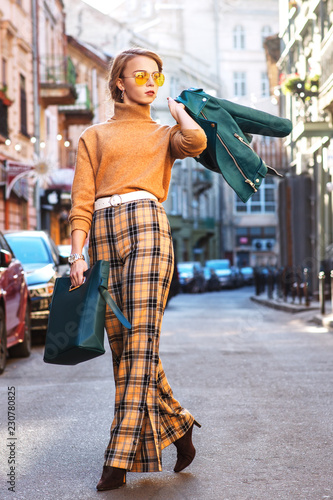 Outdoor full body fashion portrait of young fashionable girl wearing autumn outfit: glasses, turtleneck, checked culottes, heels, holding handbag, leather jacket, walking in street of european city