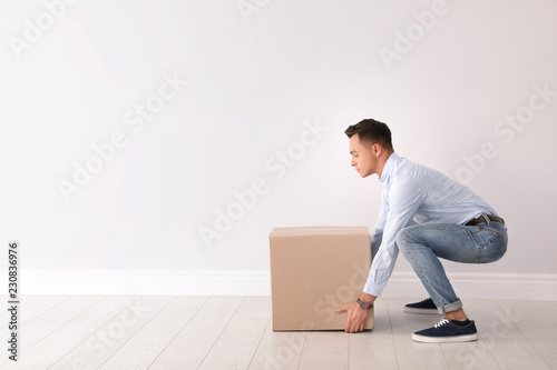 Full length portrait of young man lifting heavy cardboard box near white wall. Posture concept