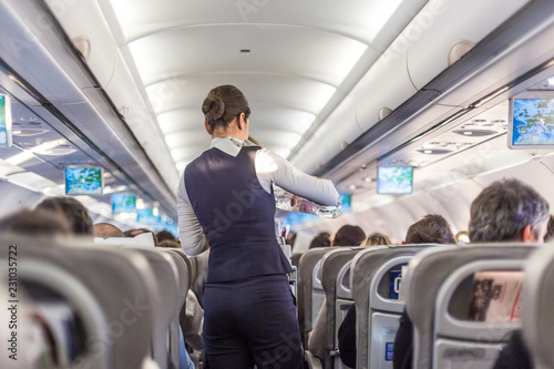 Interior of commercial airplane with flight attandant serving passengers on seats during flight. Stewardess in dark blue uniform walking the aisle. Horizontal composition.