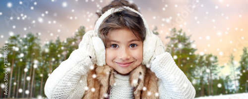 winter, people and christmas concept - happy little girl wearing earmuffs over snowy forest background