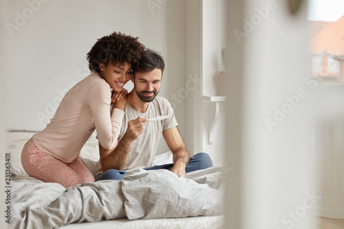 Parenthood, good news, baby expecting concept. Joyful dark skinned wife leans shoulder of her husband, look positively at pregnancy test, going to have child. Family couple on bed in modern apartment