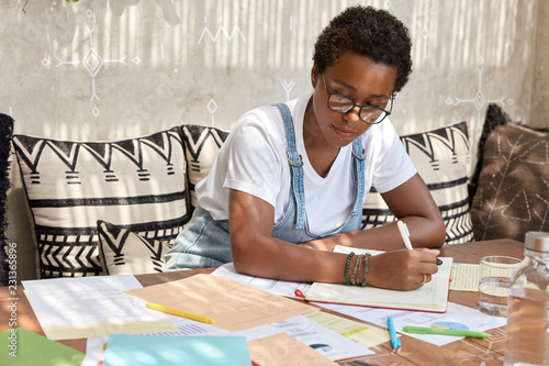 Fashionable dark skinned girl studies chart, works with many documents, writes down in notebook, has piercing, sits at desktop at comfortable couch, has serious expression, busy with autodidact