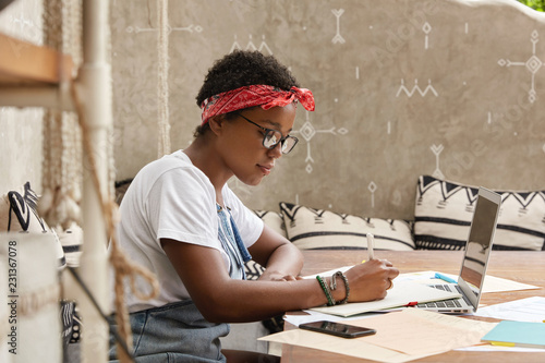 Stylish dark skinned journalist makes creative solutions, writes down some ideas on paper, surfes internet on laptop computer, prepares CV for searching new job, develops plan of effective work