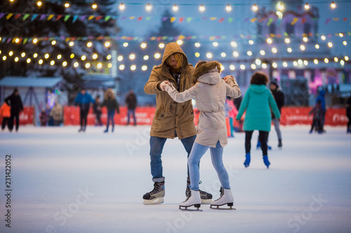 Młoda para zakochanych Kaukaski mężczyzna z blond włosami z długimi włosami i brodą i piękną kobietą bawią się, aktywnie jeżdżą na łyżwach na scenie lodowej na rynku zimą w Wigilię