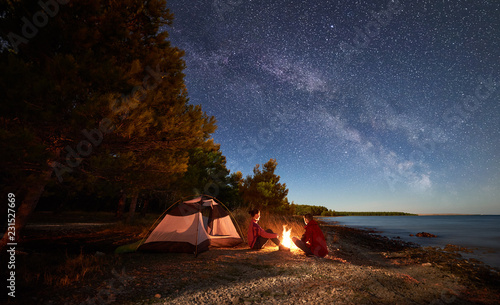 Nocne biwakowanie na brzegu. Mężczyzna i kobieta wycieczkowiczom odpoczywa przed namiotem przy ogniskiem pod wieczór niebem pełnym gwiazd i Mlecznym sposobem na błękitne wody i lasu tle. Koncepcja życia na świeżym powietrzu