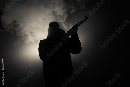 Silhouette of man with assault rifle ready to attack on dark toned foggy background or dangerous bandit in black wearing balaclava and holding gun in hand. Shooting terrorist with weapon theme decor