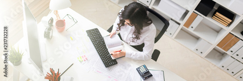 A young girl is sitting at the computer desk in the office, holding a pen in her hand and looking at the notepad.