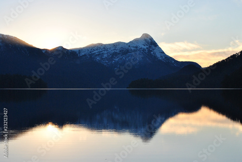 The sun hidden behind blue mountains at sunset or sunrise, with some orange glow witth clouds and clear blue sky, with clear still water with the lake in the foreground reflecting the background