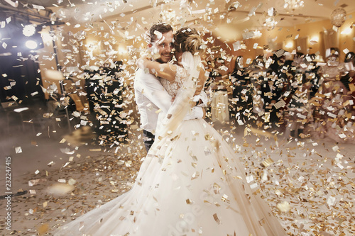 Gorgeous bride and stylish groom dancing under golden confetti at wedding reception. Happy wedding couple performing first dance in restaurant. Romantic moments