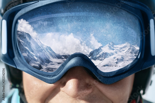 Zamyka up narciarskie gogle mężczyzna z odbiciem zaśnieżone góry. Pasmo górskie odbite w masce narciarskiej. Mężczyzna na tła niebieskim niebie. Noszenie okularów narciarskich. Sporty zimowe.