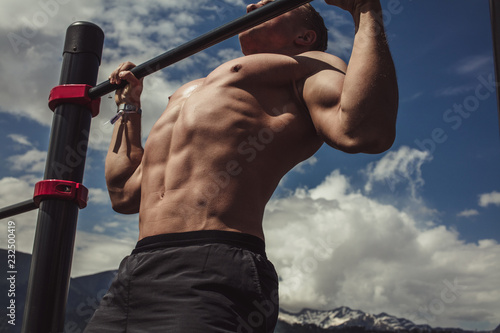 Naked torso athlete man training pull ups in amazing nature mountains landscape. Strength training fit male working out exercising outdoors in summer doing pull-ups and chin-ups on horizontal bar.
