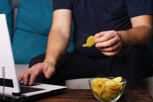 mindless snacking, overeating, lack of physical activity, laziness, homebody. fat overweight man engrossed in watching series at laptop eating fast food