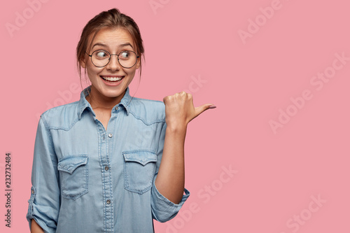 Positive Caucasian lady with toothy smile, intriguing joyful look, indicates aside, being content by something, shows free space for your advertisement or promotion, models against pink studio wall