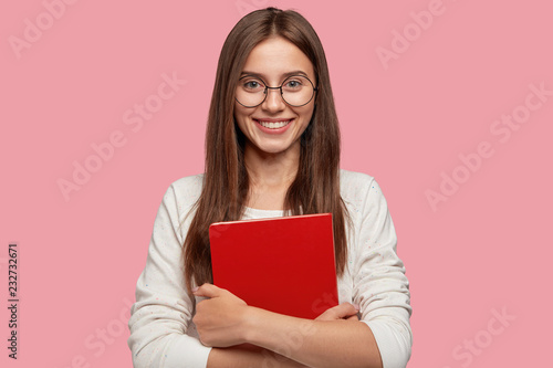 Waist up shot of pretty girl smiles pleasantly, carries red textbook closely, wears white casual jamper, models against pink background. Headshot of Caucasian lady student happy after having classes