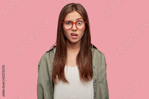 Indoor shot of indignant woman frowns face in displeasure, wears spectacles, shirt, models against pink background, doesnt like something dressed in casual shirt. Facial expressions concept.