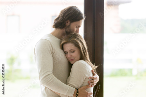 Sad young married couple embracing standing in living room opposite window at home. Sorrowful wife and husband feels unhappy, thinking about problems in relations, miscarriage, misbirth or infertility