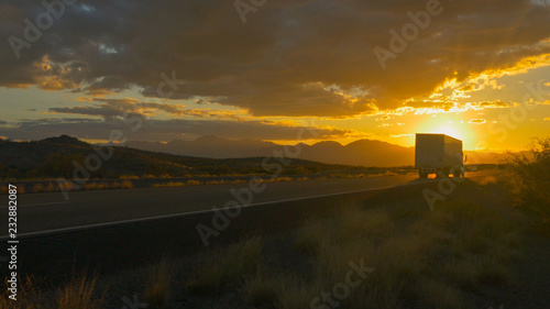 Freight semi truck speeding on empty highway over golden sun at summer sunset