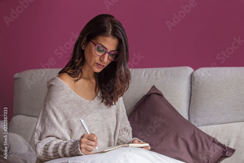 Mujer morena escribiendo a mano en el sofá en un cuaderno con bolígrafo y papel