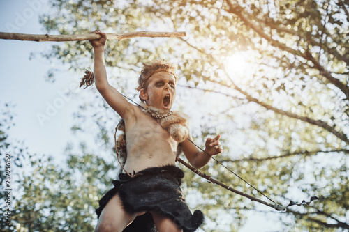 Caveman, manly boy with weapon aggressively shouting. Dramatic action photo of young primitive boy outdoors in forest. Evolution survival concept. Calm boy outside standing in attack pose. Prehistoric