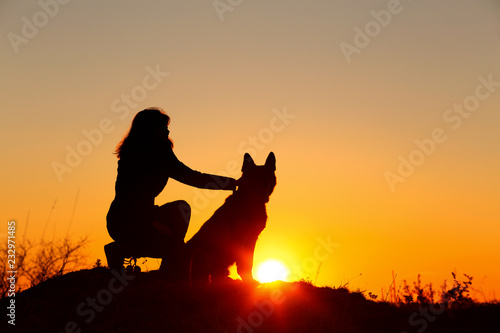 silhouette of woman embracing German Shepherd dog obediently sitting nearby, girl walking on nature with pet at sunset in a field,