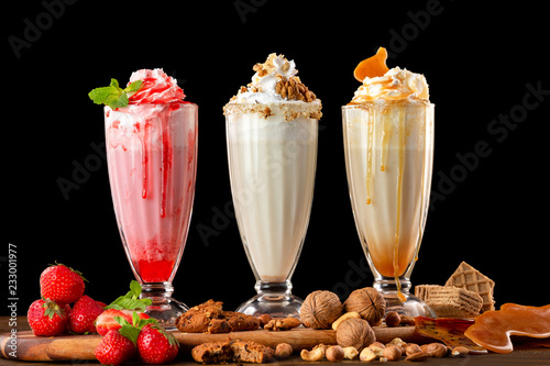 Three sweet milkshakes with nuts, caramel, strawberry and whipped cream at a wooden board on table background.